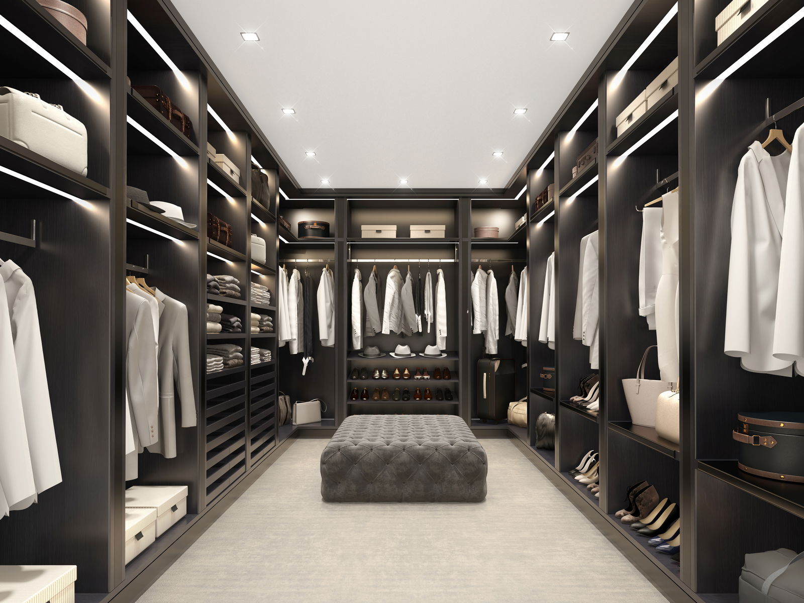 Spacious walk-in closet featuring dark wood shelves, hanging racks neatly organized with white clothing, built-in lighting, and a tufted ottoman.