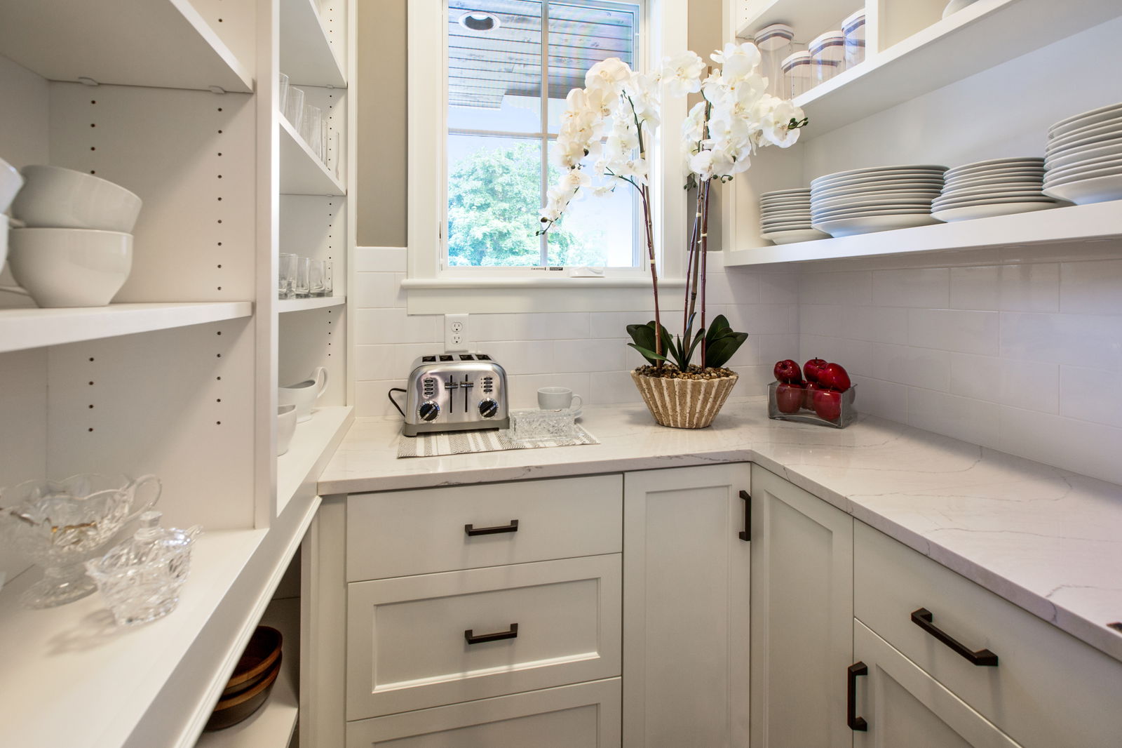 Neat pantry with white open shelves, marble countertops, shaker cabinets, dishes, glassware, a toaster, and a potted orchid under a window.