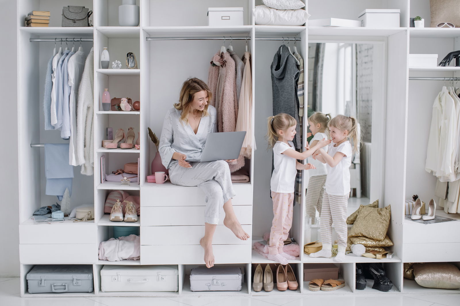Modern white closet with open shelving and hanging clothes, where a woman sits with a laptop and two young girls play near a mirror.