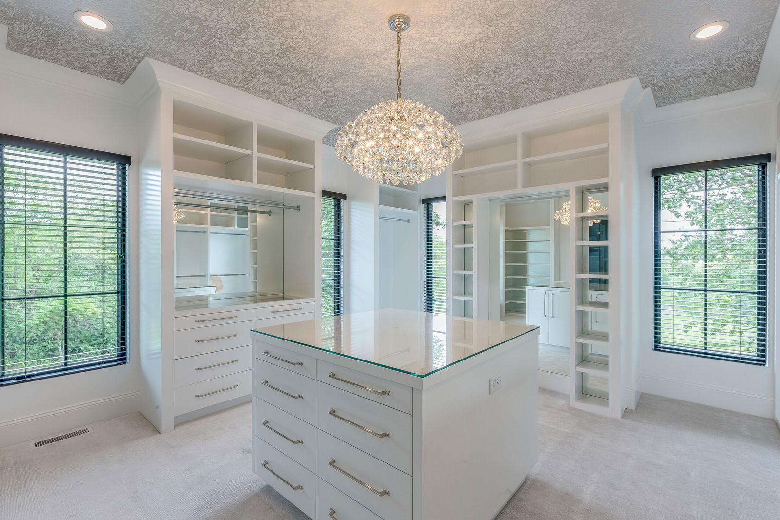 Spacious walk-in closet featuring white built-in drawers and shelves, a central island with glass top, large windows, and a crystal chandelier.