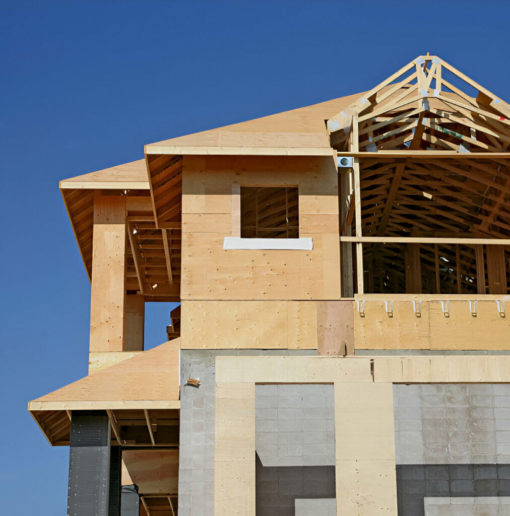 Close-up view of a two-story house under construction, showing exposed plywood sheathing, roof trusses, and concrete block foundation
