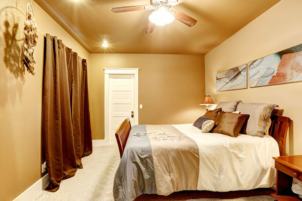 20 Inspiring Small Garage Conversion Ideas 26 Warm and inviting bedroom with earth-toned walls, a wooden bed frame, plush bedding, and decorative pillows. The room features a ceiling fan with a light fixture, rich brown curtains, and modern artwork on the wall.