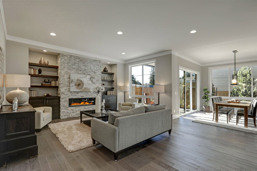Modern living room and dining area featuring a stone fireplace, grey sofa, large windows, and hardwood flooring