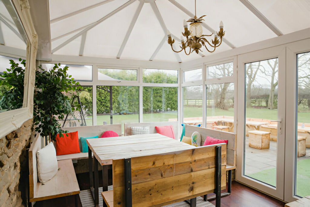 Bright and cozy sunroom with wooden furniture, colorful cushions, a chandelier, and large windows overlooking the garden