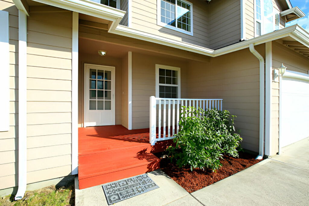 Front entrance of a modern suburban home featuring beige siding, a welcoming porch, white railings, and landscaped greenery