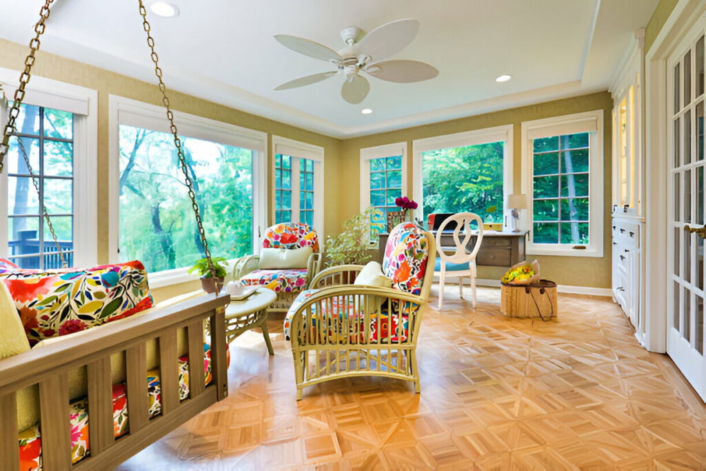 Bright sunroom with colorful floral cushions, wooden furniture, a ceiling fan, and large windows overlooking lush greenery