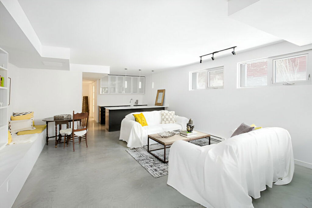 Modern finished basement living area with white sofas and kitchenette