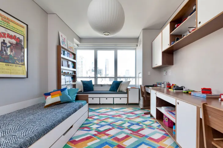 20 Inspiring Small Garage Conversion Ideas 29 A bright and colorful multipurpose kids' room featuring a bed, desk, bookshelves, and a cozy window seat with storage underneath. The room has modern white and wood furniture, a colorful geometric rug, and large windows offering a city view