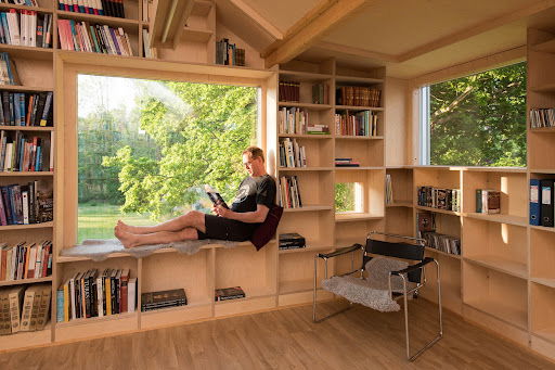 20 Inspiring Small Garage Conversion Ideas 22 A cozy home library with custom-built wooden bookshelves, a large picture window with a reading bench, and a man relaxing with a book while enjoying views of lush greenery outside.