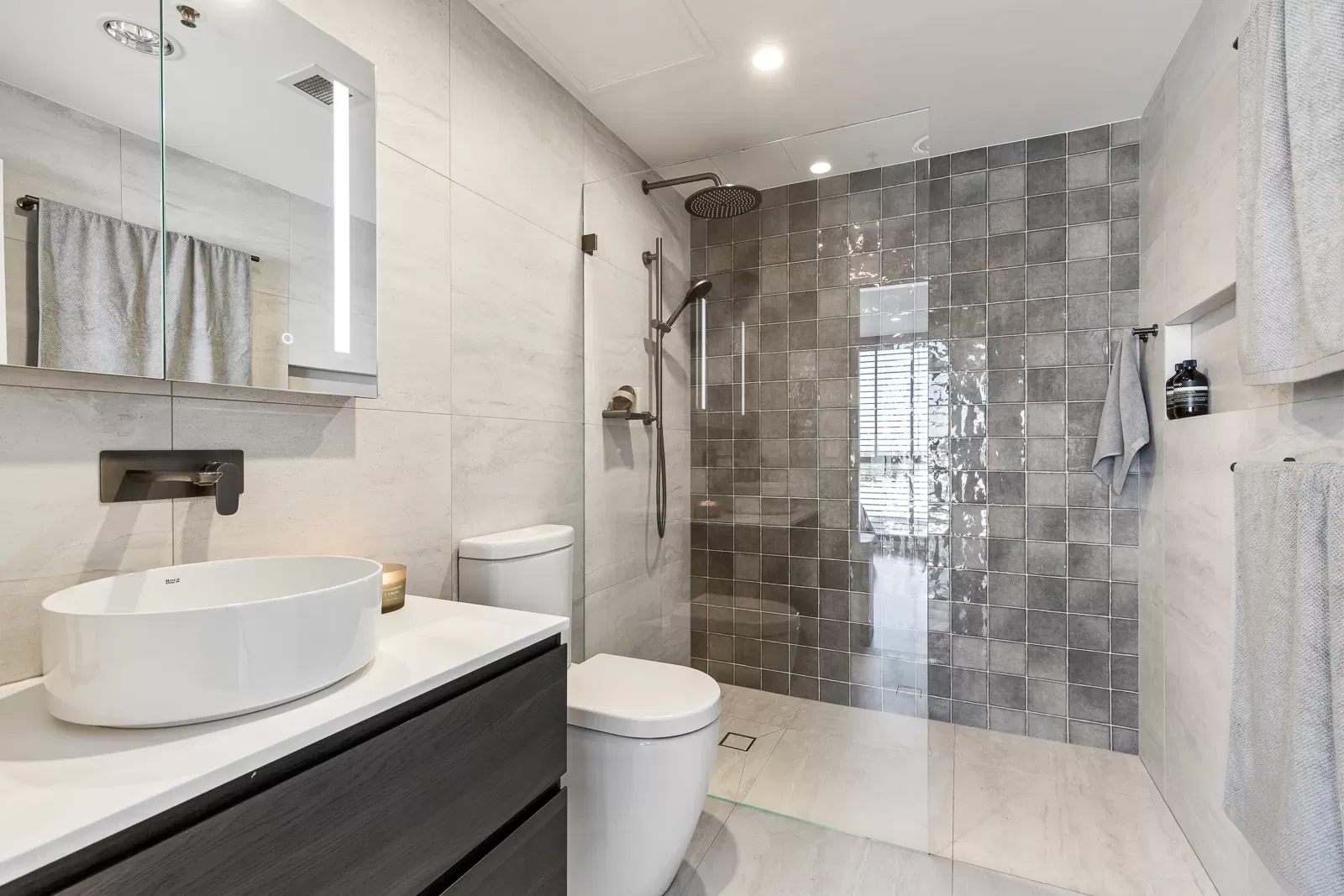 Modern bathroom with walk-in glass shower, grey tiled feature wall, floating vanity with vessel sink, and wall-mounted faucet.