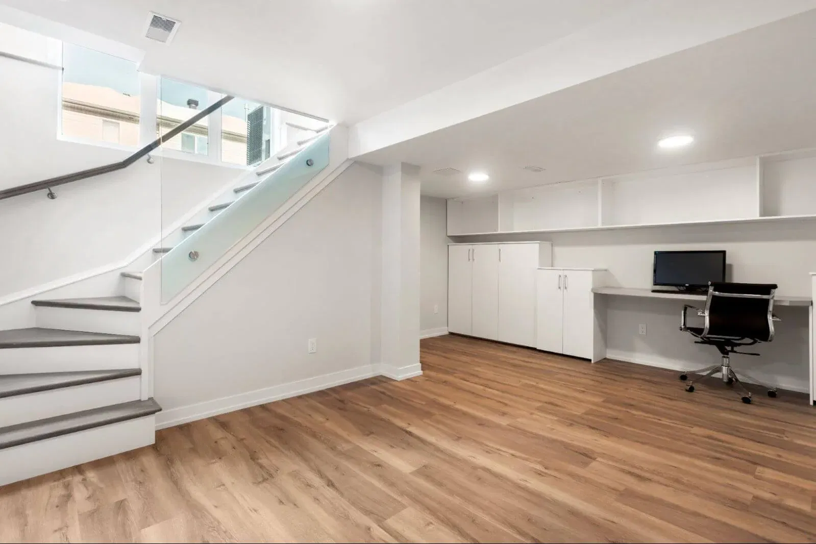 Modern finished basement with wood flooring, built-in white cabinetry, a workstation with a computer, and glass stair railing.