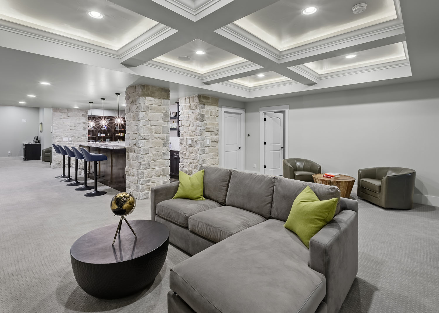 Upscale basement with coffered ceiling, stone columns, modern bar with pendant lights, and gray sectional seating.
