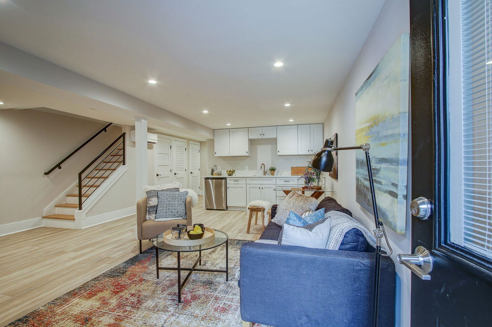 Bright and modern basement apartment with small kitchen, cozy seating area, artwork, and wood-look tile flooring.