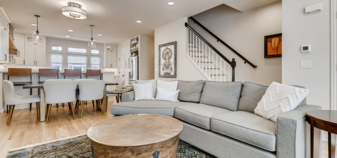 Open-concept living space with light wood floors, gray sectional sofa, rustic round coffee table, dining area, and white kitchen in the background.
