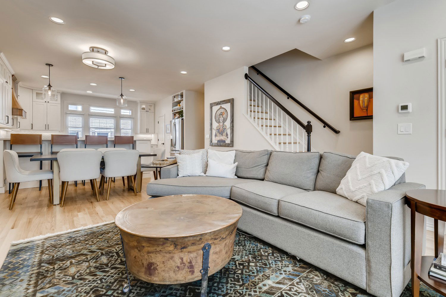 Open-concept living space with light wood floors, gray sectional sofa, rustic round coffee table, dining area, and white kitchen in the background.