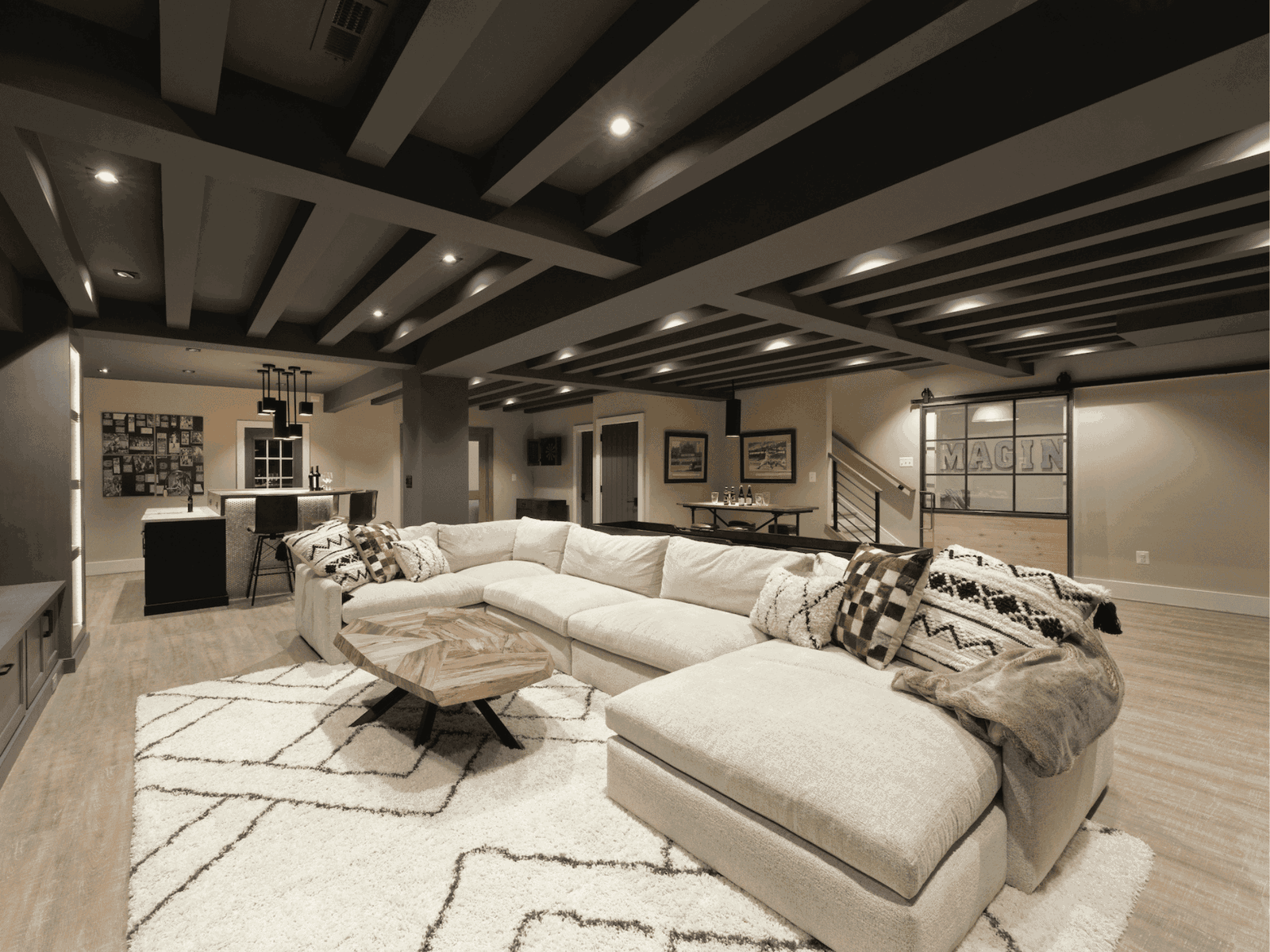 Spacious modern basement with exposed beam ceiling, oversized sectional, bar area, and warm neutral tones.