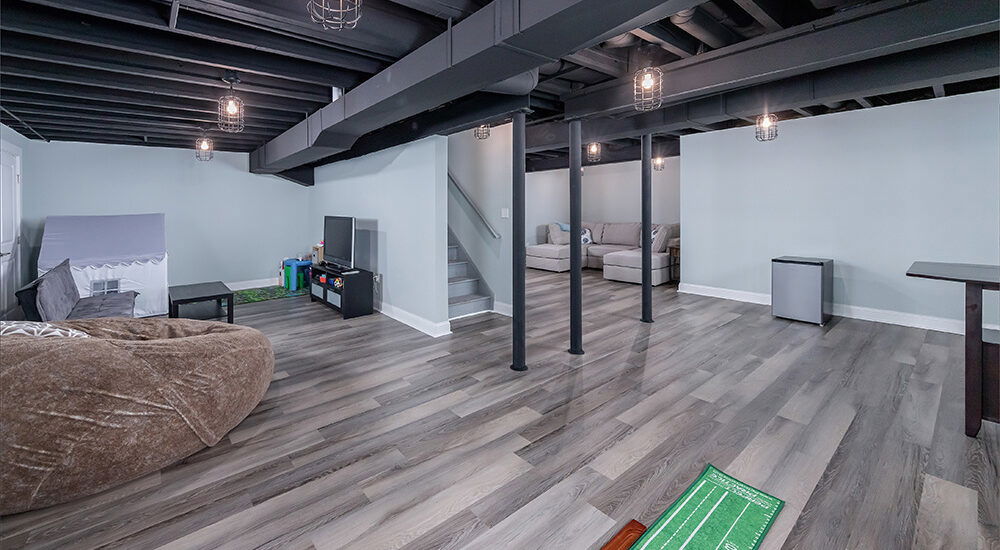 Open-concept basement with exposed black ceiling beams, vinyl plank flooring, lounge area, kids’ play zone, and putting green.