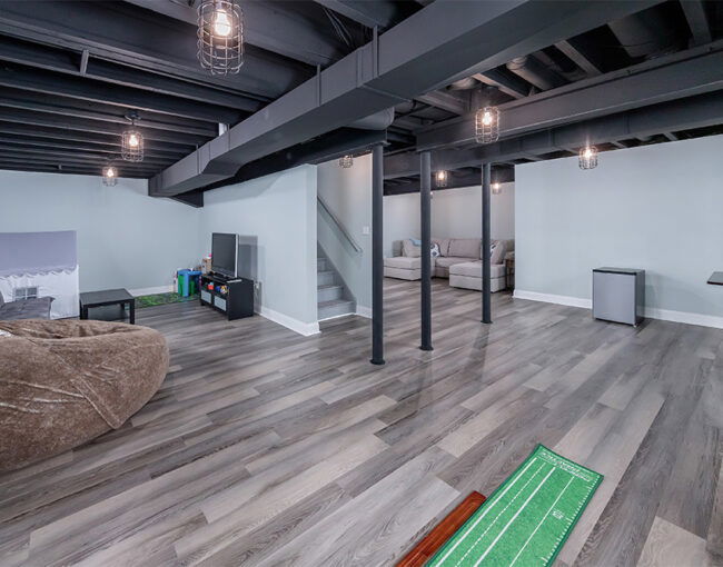 Open-concept basement with exposed black ceiling beams, vinyl plank flooring, lounge area, kids’ play zone, and putting green.