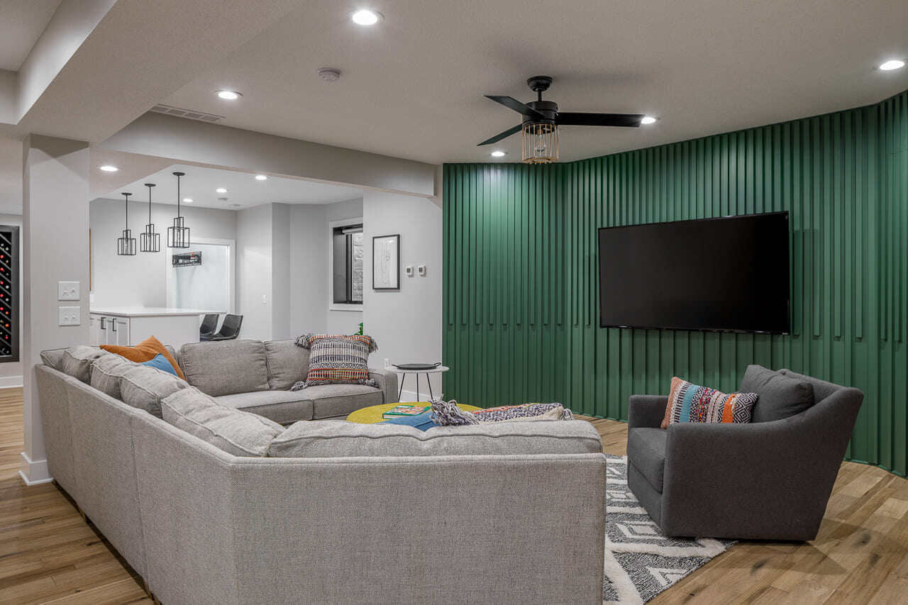 Modern basement entertainment room with sectional sofa, green accent wall, mounted TV, and open-concept bar area.