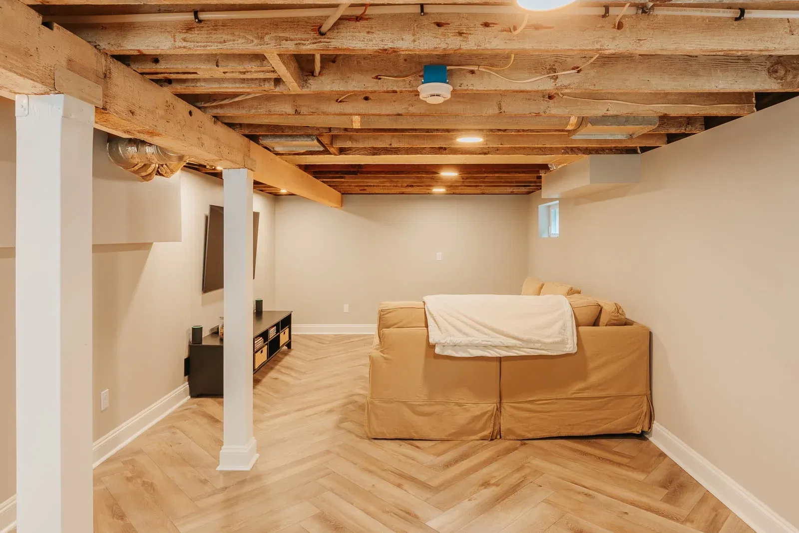 Cozy basement with exposed wooden beam ceiling, tan sectional sofa, herringbone wood flooring, and minimalist TV setup.