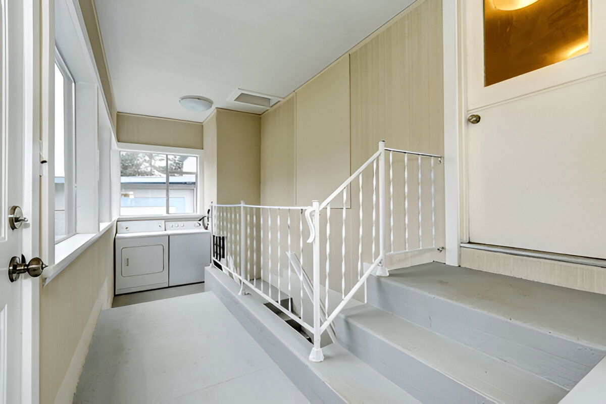 Simple laundry area with washer and dryer, beige panel walls, white stair railing, and large windows for natural light.