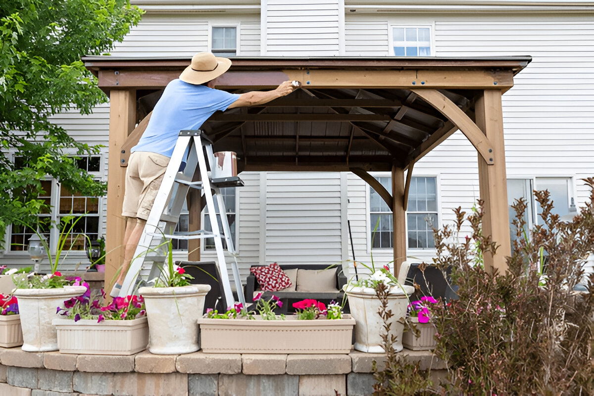 Person staining a wooden gazebo in a backyard using a ladder and paintbrush