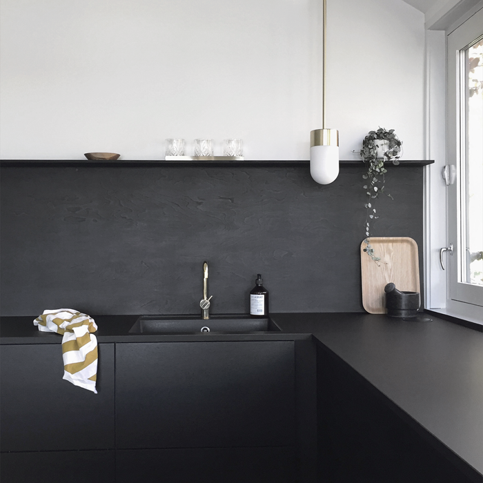 Minimalist kitchen with matte black cabinets and backsplash, gold faucet, white pendant light, and modern decor elements.