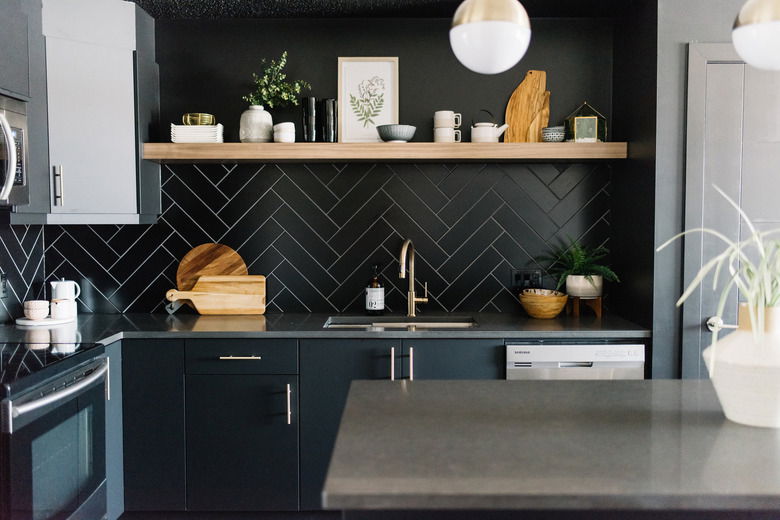 Contemporary kitchen with matte black cabinets, herringbone black tile backsplash, wooden floating shelf with decor, and brushed gold faucet.
