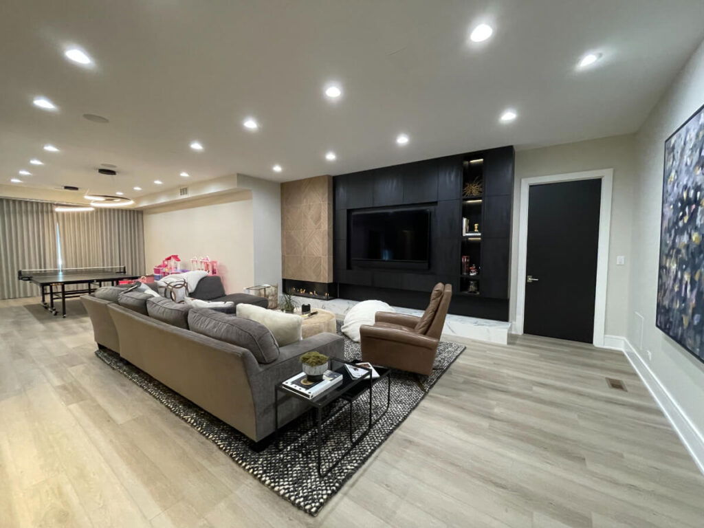 Modern basement with sectional sofa, accent chairs, large wall-mounted TV, black built-in media wall, and ping pong table in the background.