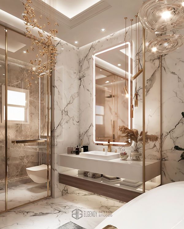 Luxury marble bathroom with gold accents, illuminated mirror, floating vanity, and glass-enclosed shower.