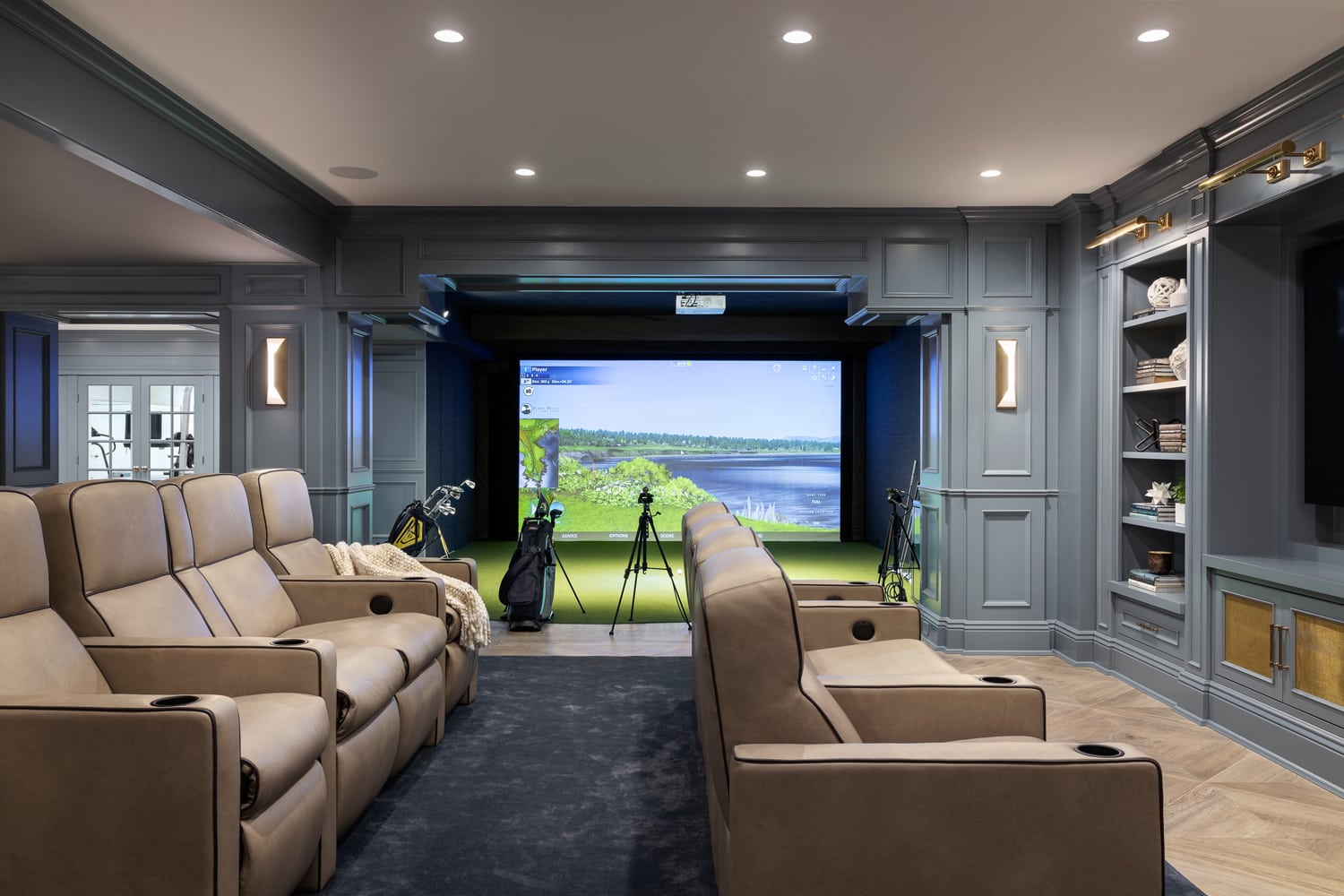 Elegant basement media room with tan recliners and an immersive golf simulator setup, surrounded by custom built-in cabinetry.