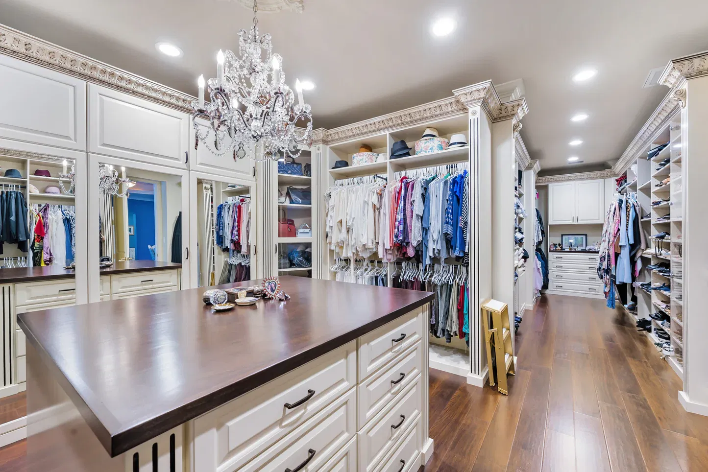  A spacious and elegantly designed walk-in closet featuring a crystal chandelier, built-in shelves, hanging rods, and a central island with drawers
