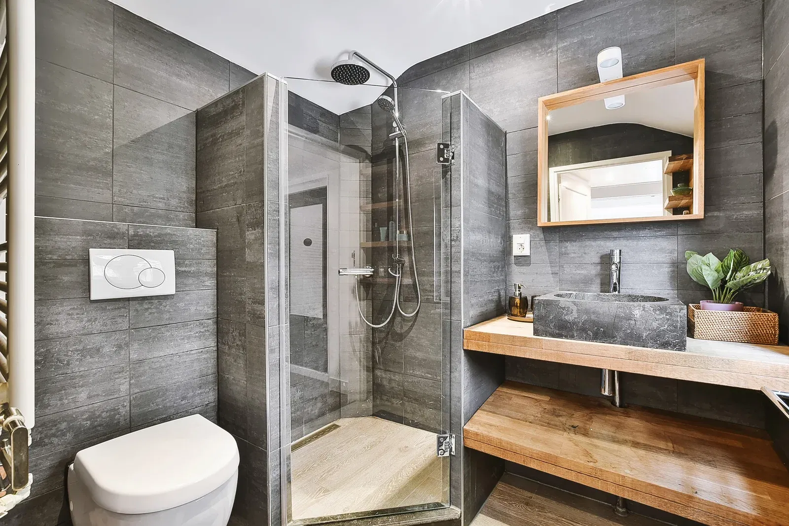 Modern small bathroom with dark grey tiles, glass corner shower, wooden vanity, and stone sink.