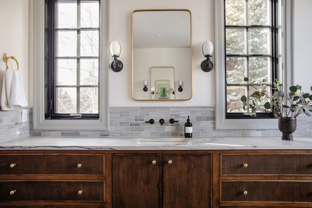 Classic bathroom vanity with dark wood cabinetry, marble countertop, gold-framed mirror, and double windows