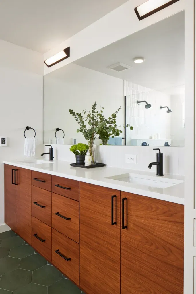 Contemporary bathroom featuring a wooden double vanity with white quartz countertop, dual sinks, black faucets and handles, wall-mounted mirror, and indoor plants for a fresh touch.