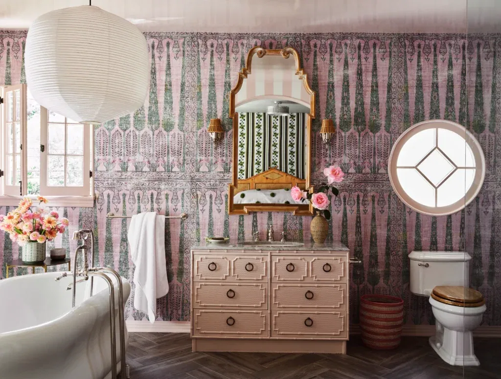 Luxurious bathroom with patterned pink wallpaper, ornate pink vanity, sculptural mirror, and circular window creating a romantic vintage vibe.