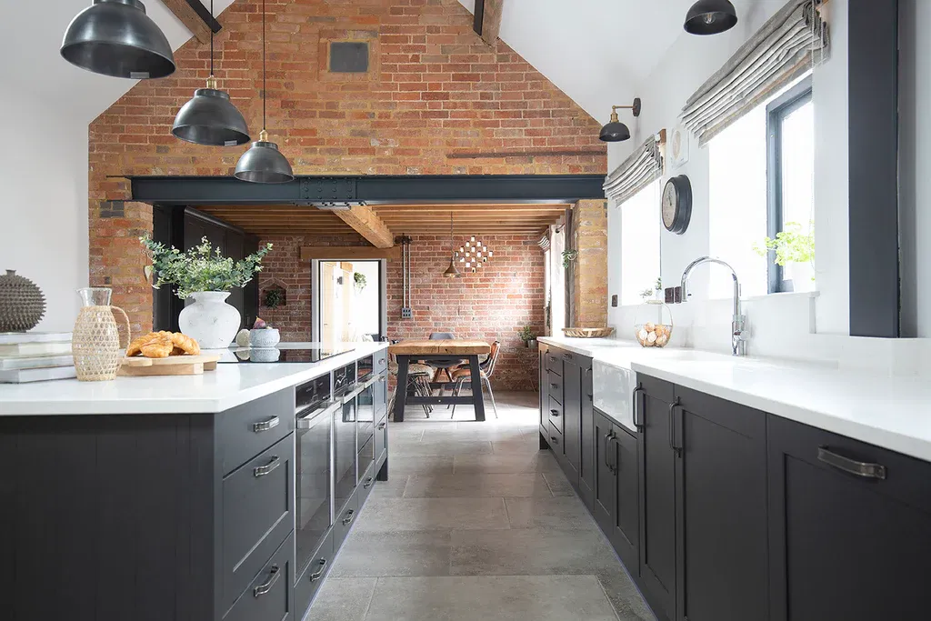 Industrial-style kitchen featuring grey tumbled porcelain stone floors, matte black cabinetry, white countertops, and exposed red brick walls with black-framed windows and industrial pendant lighting.