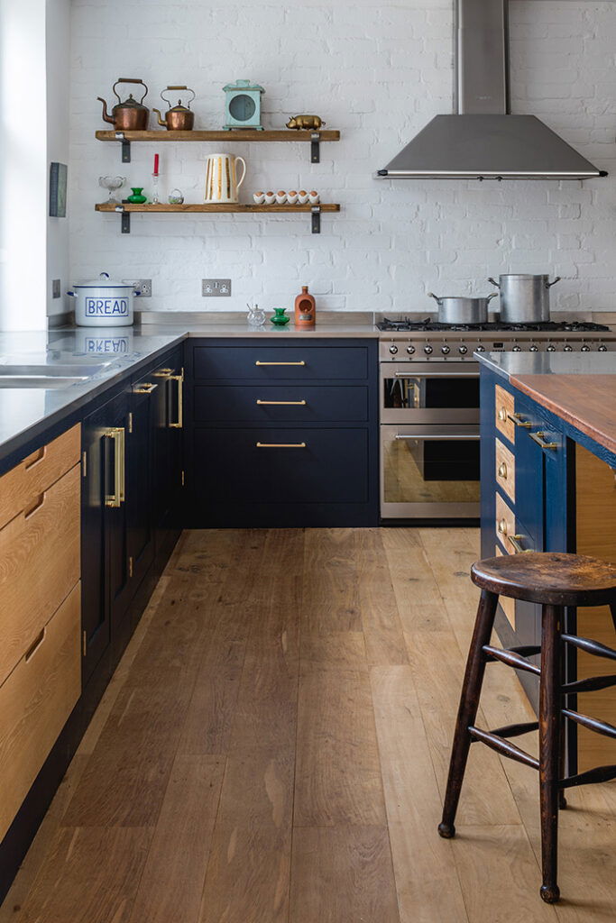 Rustic kitchen with Victorian beam oak wood flooring, navy and natural wood cabinetry, brass hardware, and exposed white brick walls.