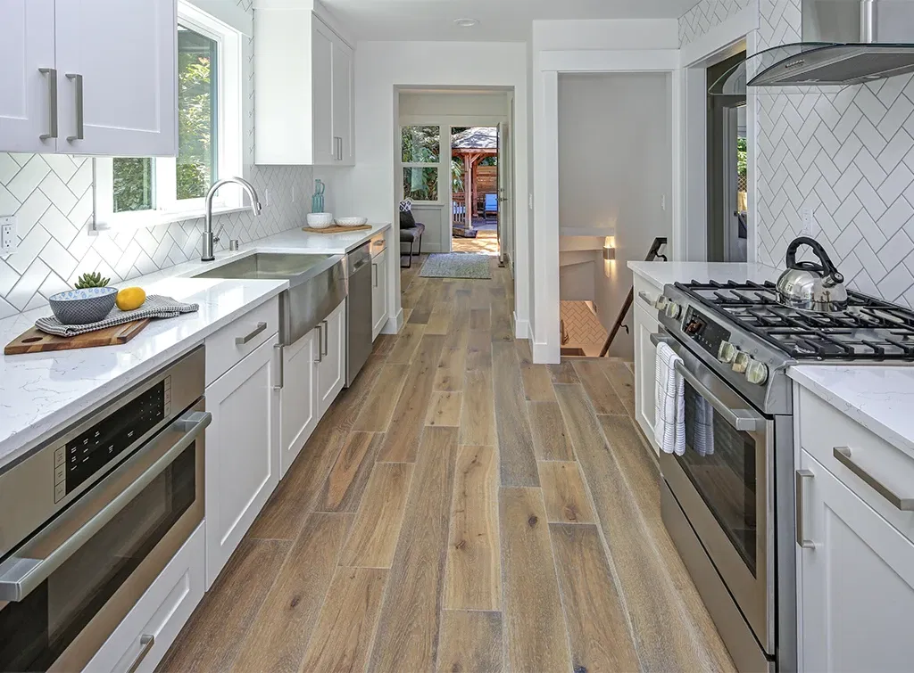 Contemporary galley kitchen with white cabinetry, stainless steel appliances, and wood-look tile flooring in a natural finish.