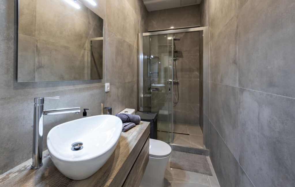 Modern small bathroom with gray large-format tiles, floating wooden vanity, white vessel sink, wall mirror, and a glass-enclosed shower.