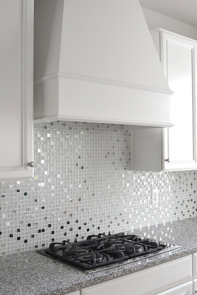 Close-up of a white kitchen featuring a glass and metal mosaic backsplash and a gas stovetop