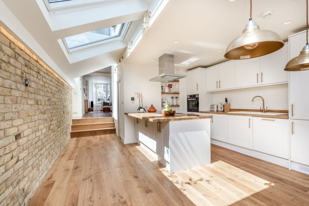 Modern white kitchen with wood countertops, island, oak flooring, exposed brick wall, and skylights bringing in natural light.