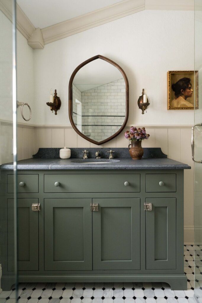 Classic bathroom vanity with sage green cabinets, arched wood mirror, dark countertop, and vintage wall sconces for an antique aesthetic.