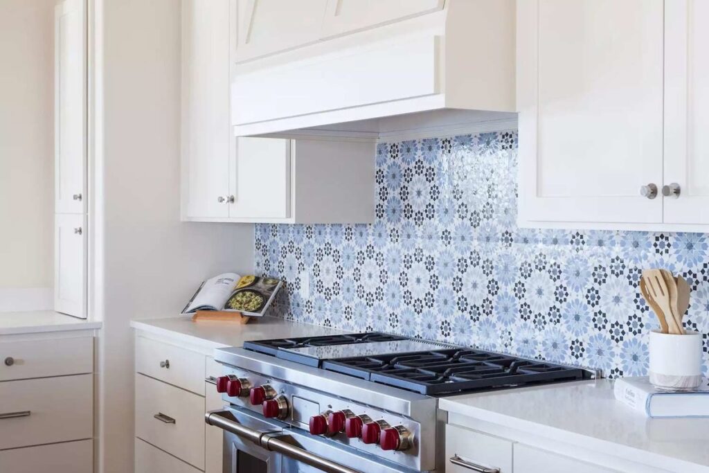 Modern white kitchen featuring a patterned blue Moroccan tile backsplash, stainless steel stove with red knobs, and white cabinetry