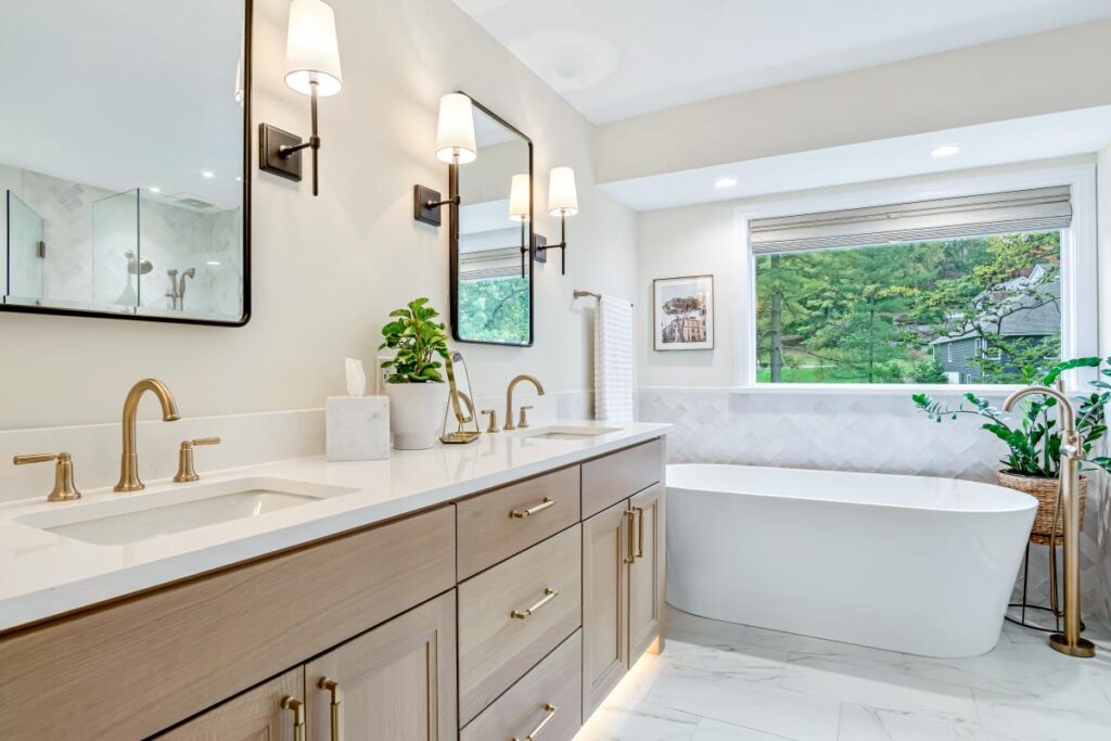 Modern bathroom featuring a double vanity, gold fixtures, freestanding tub, large window with greenery views, and biophilic design elements.
