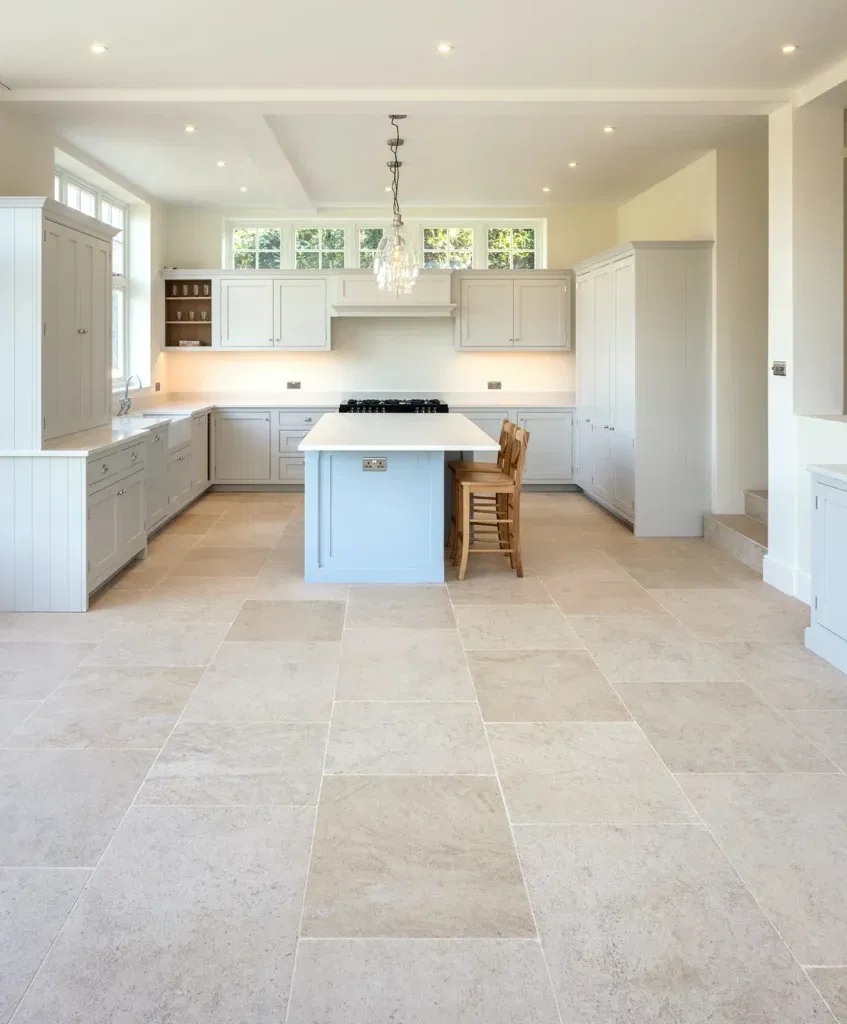 Spacious kitchen with light beige limestone tile flooring, pastel gray cabinets, central island with wooden stools, and modern lighting