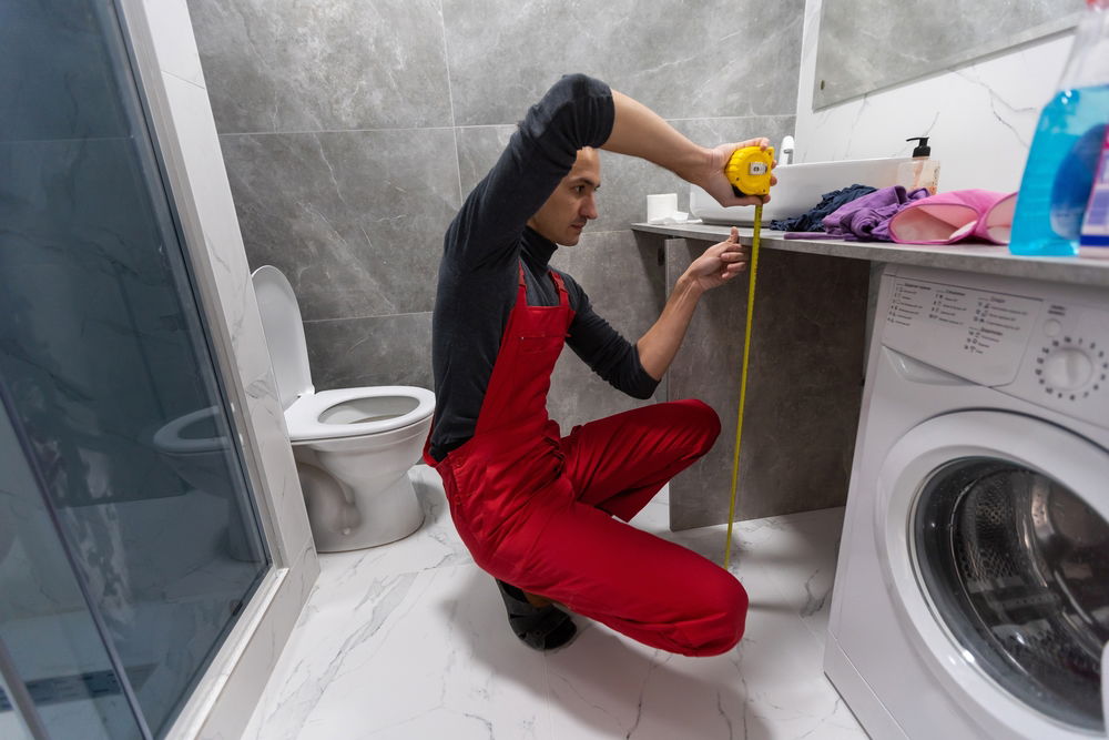 The image shows a man in a red jumpsuit and black gloves crouching in a bathroom, holding a tape measure and inspecting a fixture or appliance.