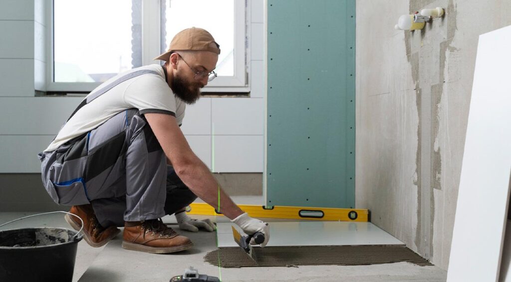 A construction worker in overalls and gloves installing a large floor tile in a partially renovated bathroom, using adhesive and a notched trowel.